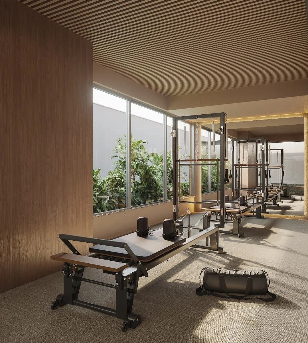 Fotografia do fitness center do Ipanema Almar, com equipamentos de pilates modernos, iluminação natural abundante através de amplas janelas com vista para vegetação exuberante, piso em carpete claro e teto em madeira ripada.